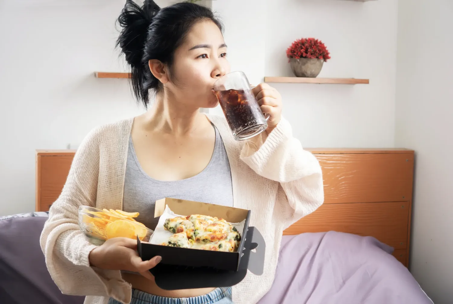 A woman sitting on her bed in the evening with a tray of food and a cold drink, eating while looking away from the food.